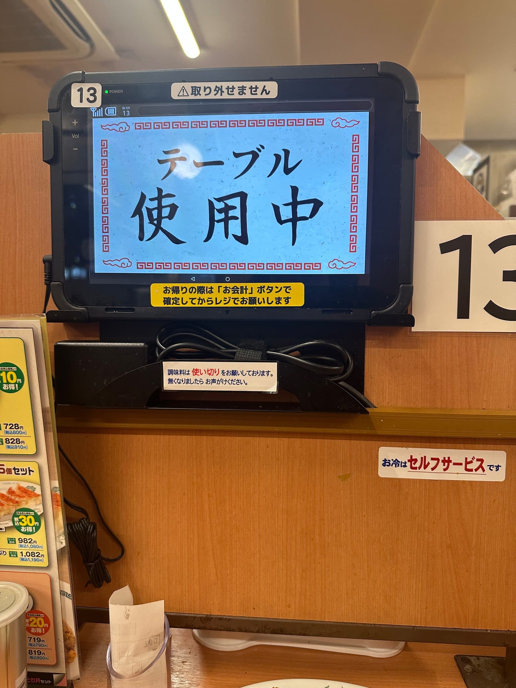 En fastmonterad pekplatta vid ett restaurangbord i Japan visar en blå skärm med japansk text som anger att bordet är upptaget. Plattan sitter på en träskiljevägg med bordsnumret 13 bredvid och en menykartong till vänster.