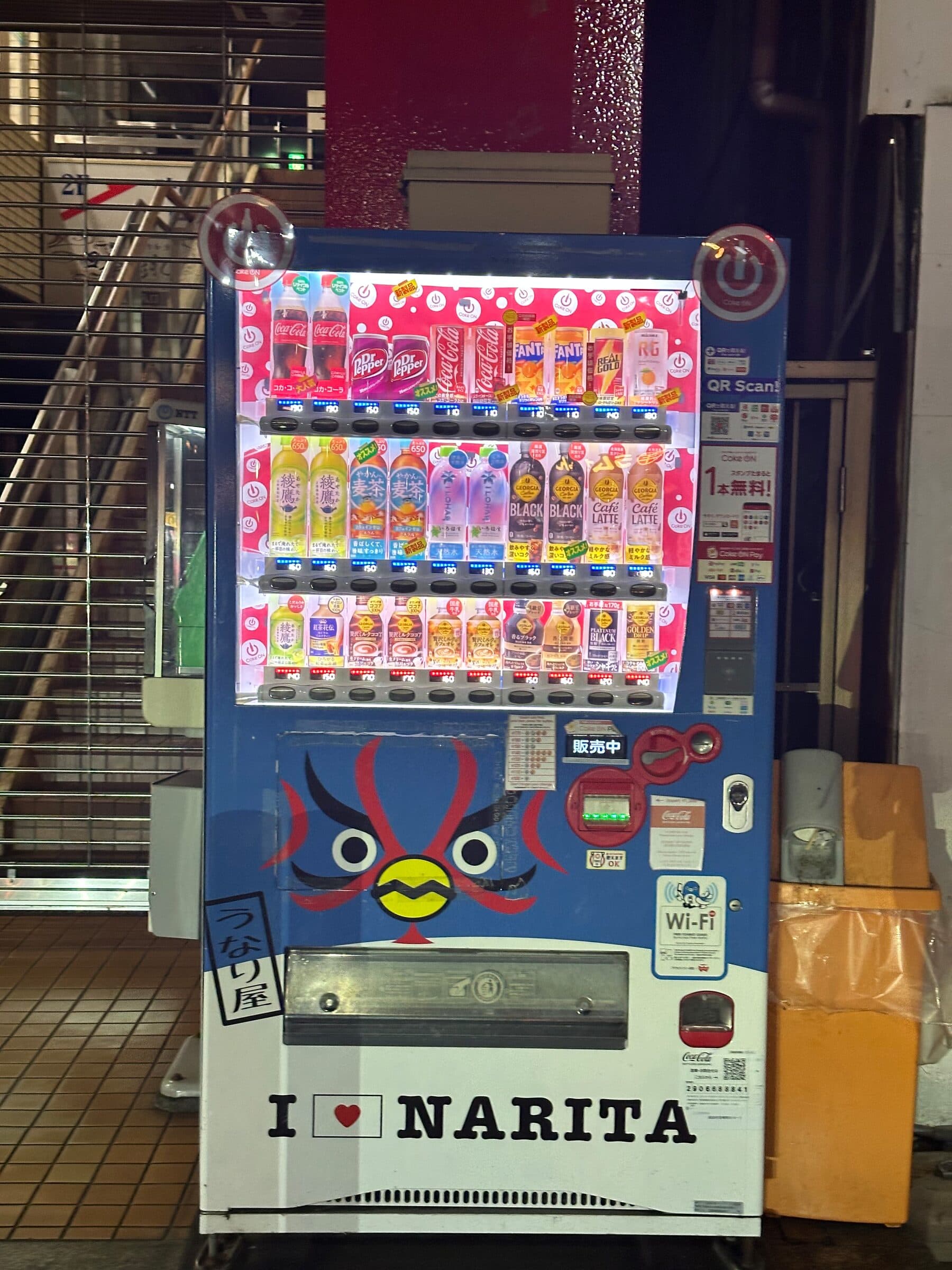 Dryckesautomat på en gata i Narita nattetid, dekorerad med en illustrerad festivalmask i rött och blått på nedre fronten och texten I love Narita. De upplysta hyllorna visar läsk och flaskor, och en skylt intill uppmanar till betalning med QR-kod.