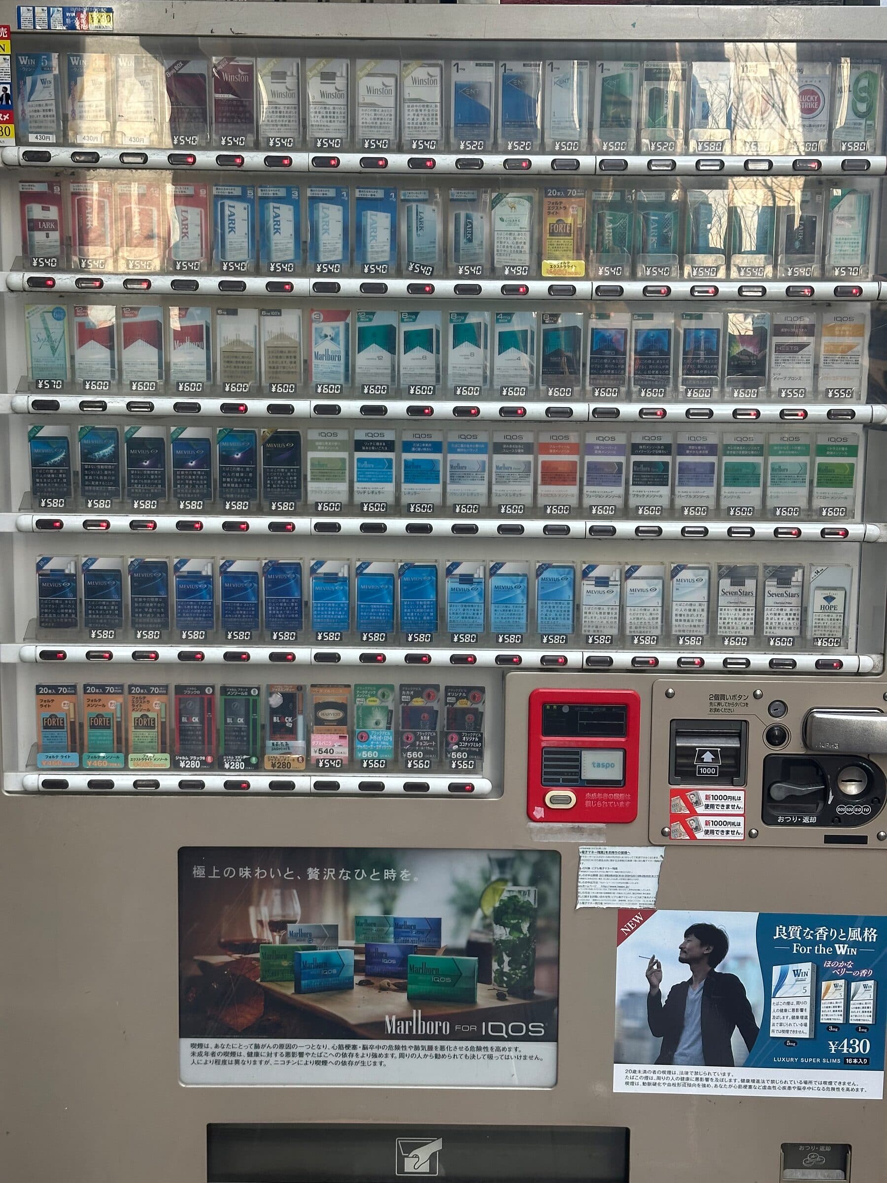 Närbild på fronten av en japansk tobaksautomat i Tokyo. Bakom glaset står cigarettpaket i sex rader med prislappar i yen, och till höger sitter en röd taspo-läsare för åldersverifiering intill sedelinkast och myntinkast.