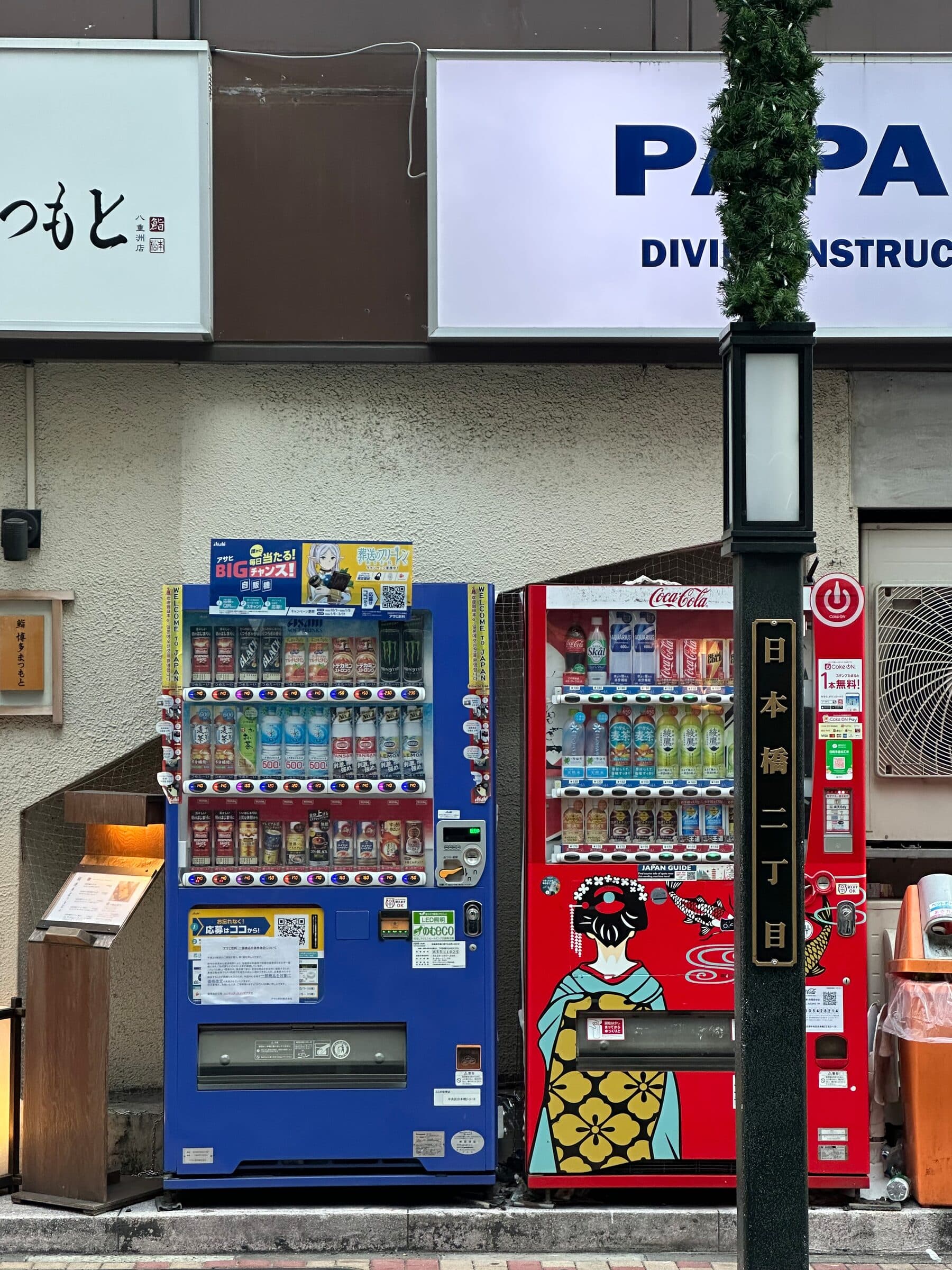 En blå och en röd dryckesautomat står tätt intill varandra mot en husvägg i stadsdelen Nihonbashi i Tokyo. Den röda automaten är dekorerad med en illustration av en geisha i kimono, och en gatlykta med gatunamnsskylt står framför.