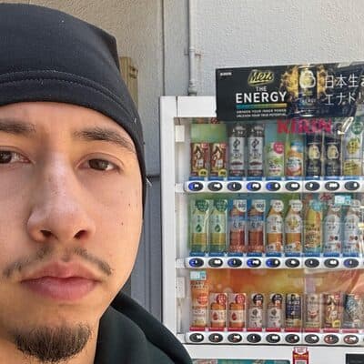 Eric framför en dryckesautomat på en gata i Japan. Han bär mössa och mörk jacka, och bakom honom syns automatens upplysta hyllor med flaskor och burkar.