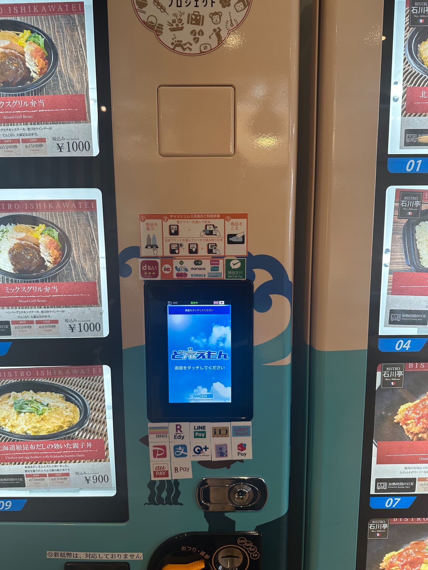 Matautomat i Japan med fotograferade menypaneler i två spalter längs sidorna. Varje panel visar en färdig rätt med namn, nummer och pris i yen, och i mitten sitter en pekskärm med en rad dekaler för olika betalappar över och under.