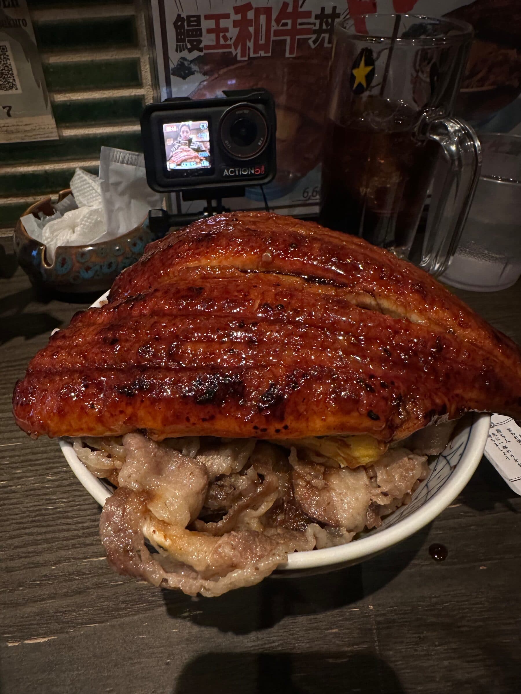 En stor skål med en glaserad grillad ålfilé som täcker hela skålens öppning, serverad över kött och ris. Bakom skålen står en actionkamera på stativ riktad mot maten och ett glas med dryck.