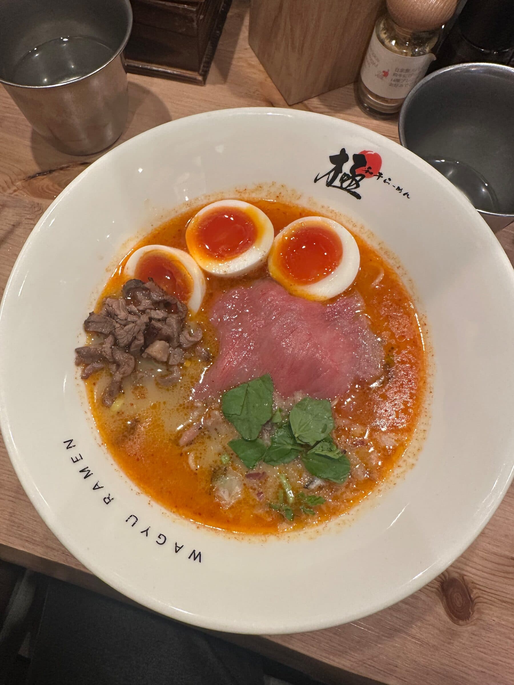En vit skål med orangeröd ramenbuljong, två halverade kokta ägg med lös gula, en tunn skiva rått nötkött, hackad svamp och färska örter. På skålens kant står texten wagyu ramen.