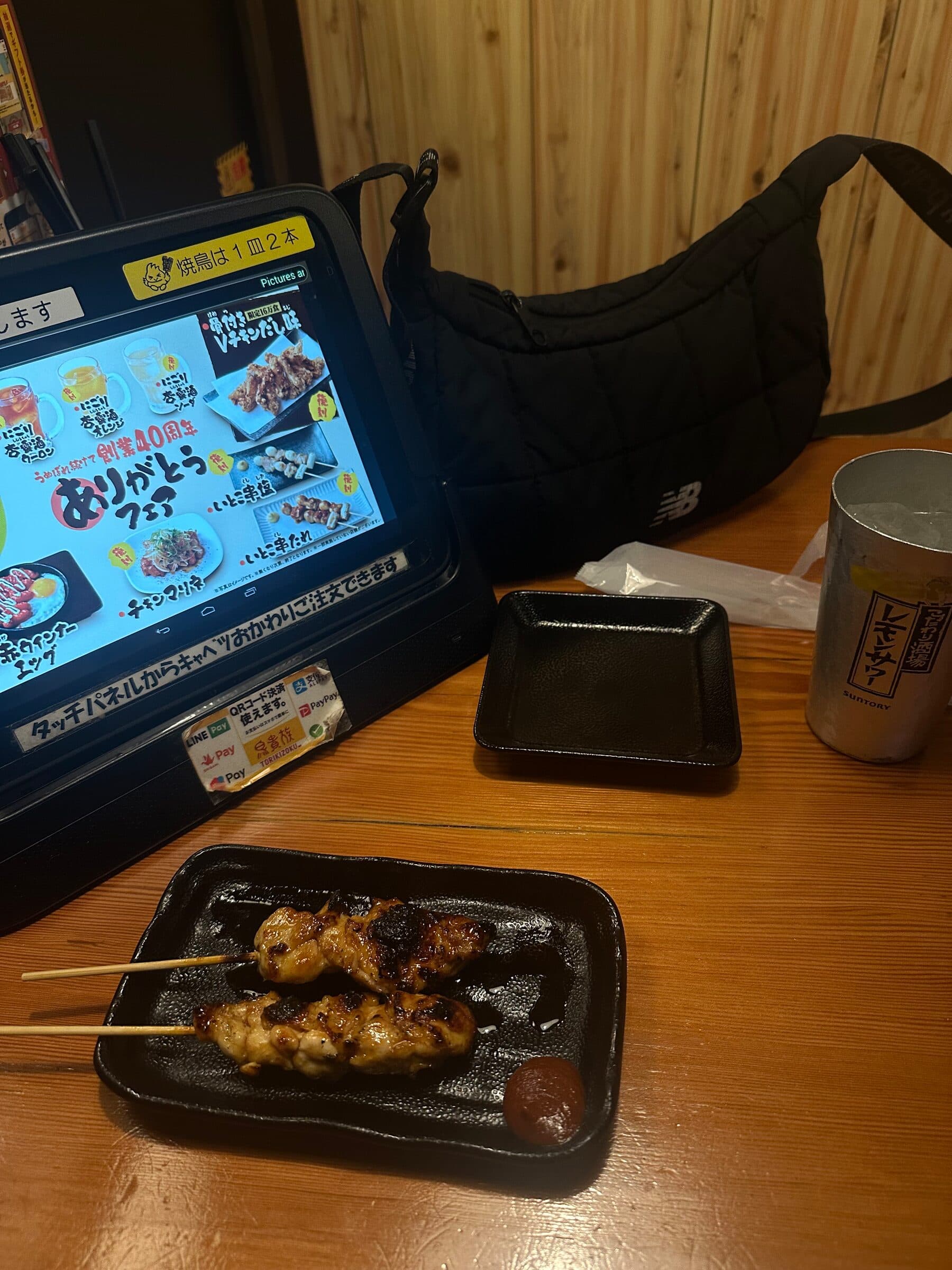 Ett träbord på en japansk restaurang med en fastmonterad beställningsplatta till vänster som visar menybilder, och framför den ett svart fat med två grillade spett och en kopp te.