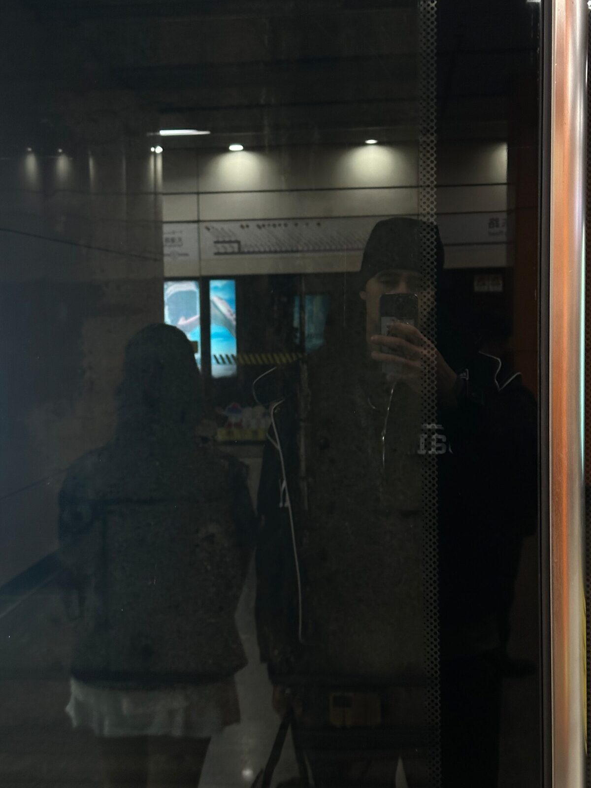 Eric speglar sig i en mörk glasruta i Kina medan han fotograferar med telefonen. Bakom glaset skymtar en upplyst lokal med skyltar i taket, och ytterligare en person syns till vänster i reflektionen.